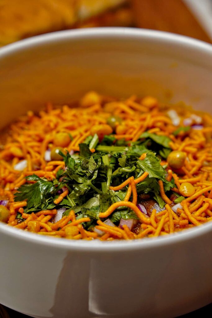 An extreme close-up of a bowl of Misal, highlighting the texture of the crunchy yellow farsan, green cilantro, and spicy red curry.