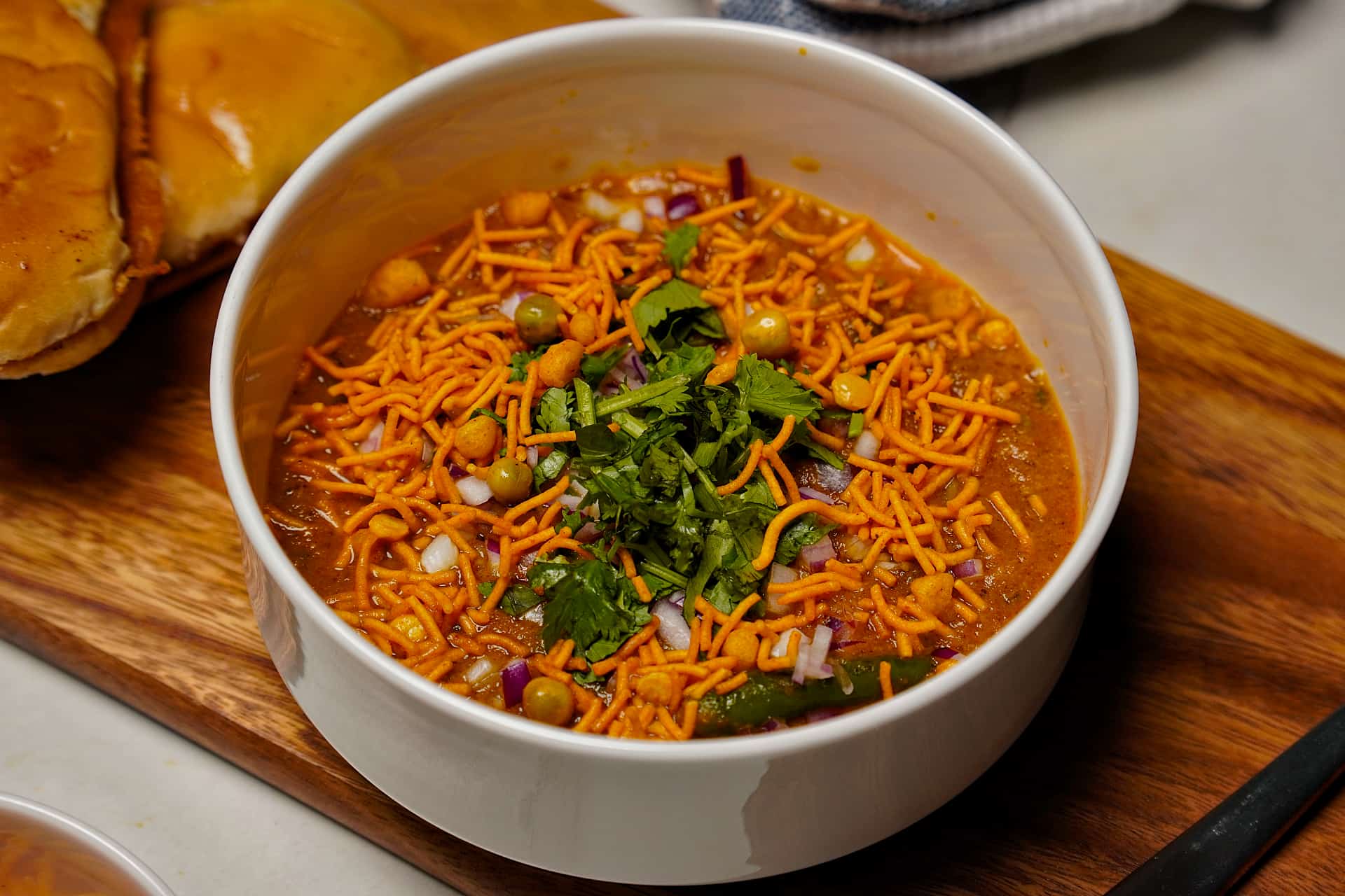 A high-angle shot of a white bowl filled with spicy Misal topped with farsan, chopped onions, and fresh cilantro, served on a wooden board.