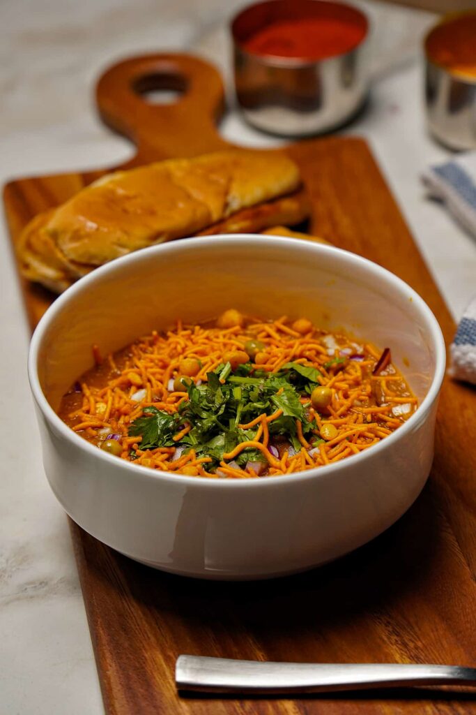 A wide-angle view of a bowl of spicy Misal served with buttered pav bread and small bowls of spices in the background.