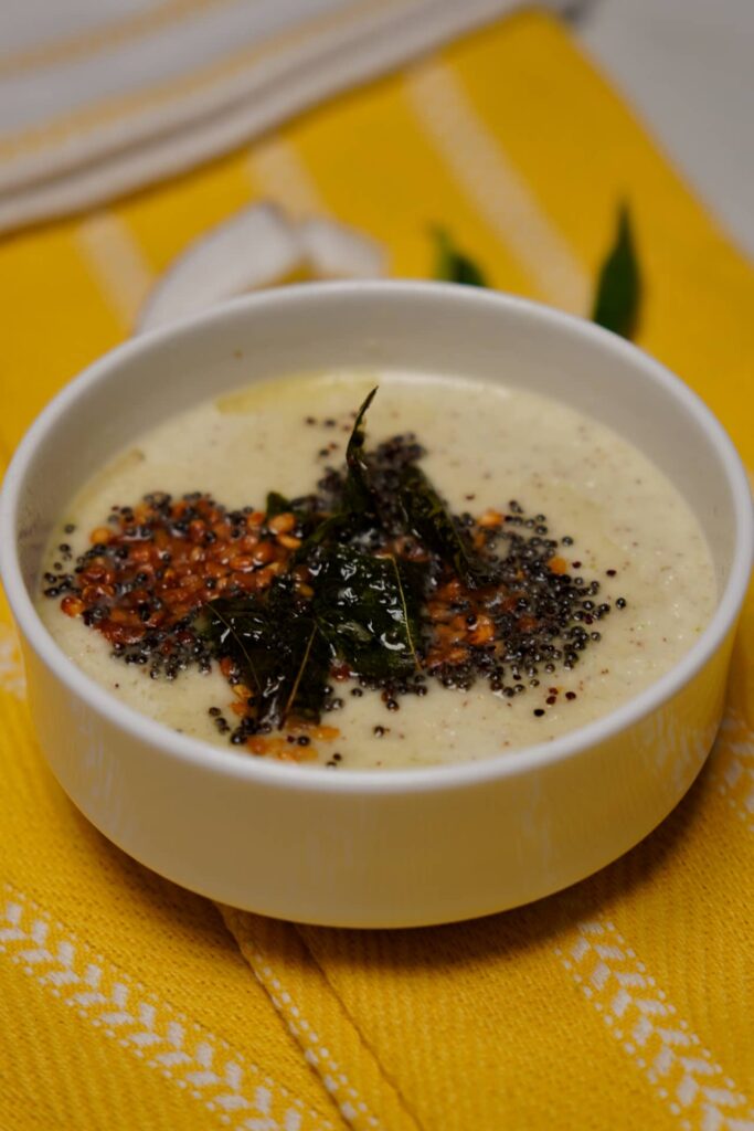 Side view of a bowl of coconut chutney featuring fresh coconut slices and green chilies in the background, styled on a yellow cloth.