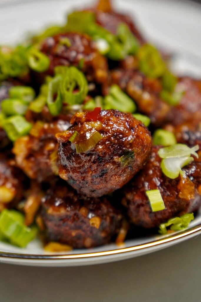 Extreme close-up of a single crispy Manchurian ball coated in a glossy, savory brown sauce with bits of minced garlic and chili.