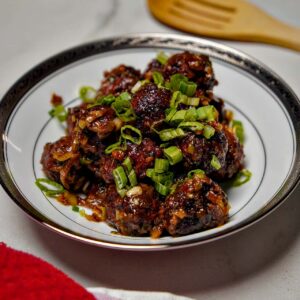 A wide-angle shot of a bowl of Veg Manchurian on a marble countertop with a wooden spatula and a red kitchen towel in the background.