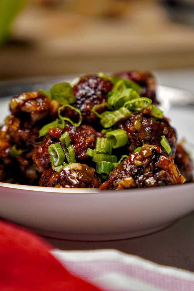Low-angle side view of a heap of Manchurian balls in a decorative bowl, highlighting the texture of the sauce and the garnish.