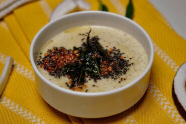 A white bowl of creamy coconut chutney topped with a tempering of black mustard seeds, red dal, and fried curry leaves on a yellow textured napkin.