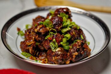 A white bowl filled with dark, glazed Indo-Chinese Veg Manchurian balls topped with a generous amount of freshly chopped green onions.