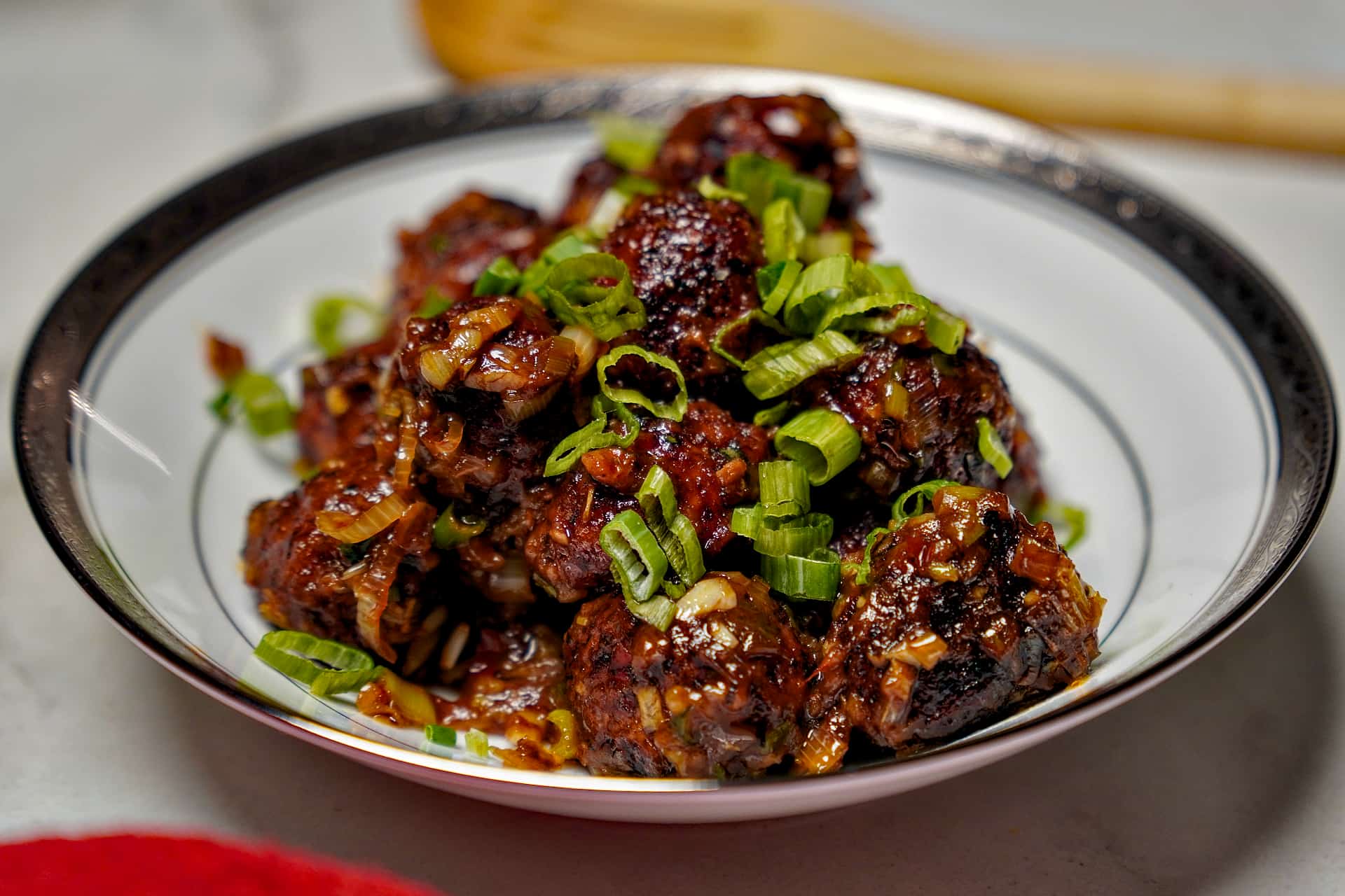 A white bowl filled with dark, glazed Indo-Chinese Veg Manchurian balls topped with a generous amount of freshly chopped green onions.