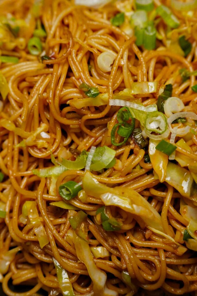 Extreme close-up of glossy, soy-sauce coated noodles mixed with vibrant green spring onions and sautéed cabbage.