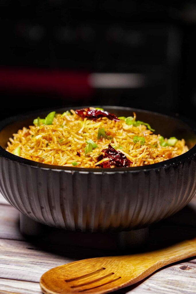 A low-angle profile shot of a full bowl of spicy Schezwan rice with a wooden spatula in the foreground, perfect for a blog header.