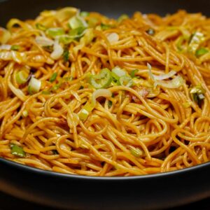 A close-up side view of a steaming heap of vegetable noodles in a dark bowl, highlighting the colorful vegetable garnishes.