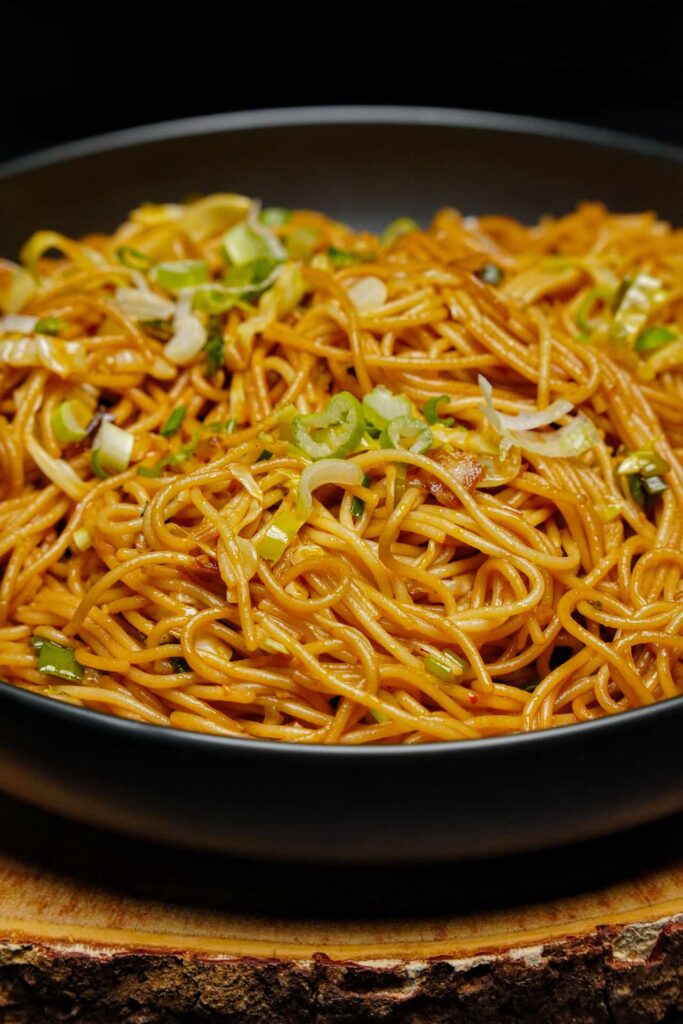 A close-up side view of a steaming heap of vegetable noodles in a dark bowl, highlighting the colorful vegetable garnishes.