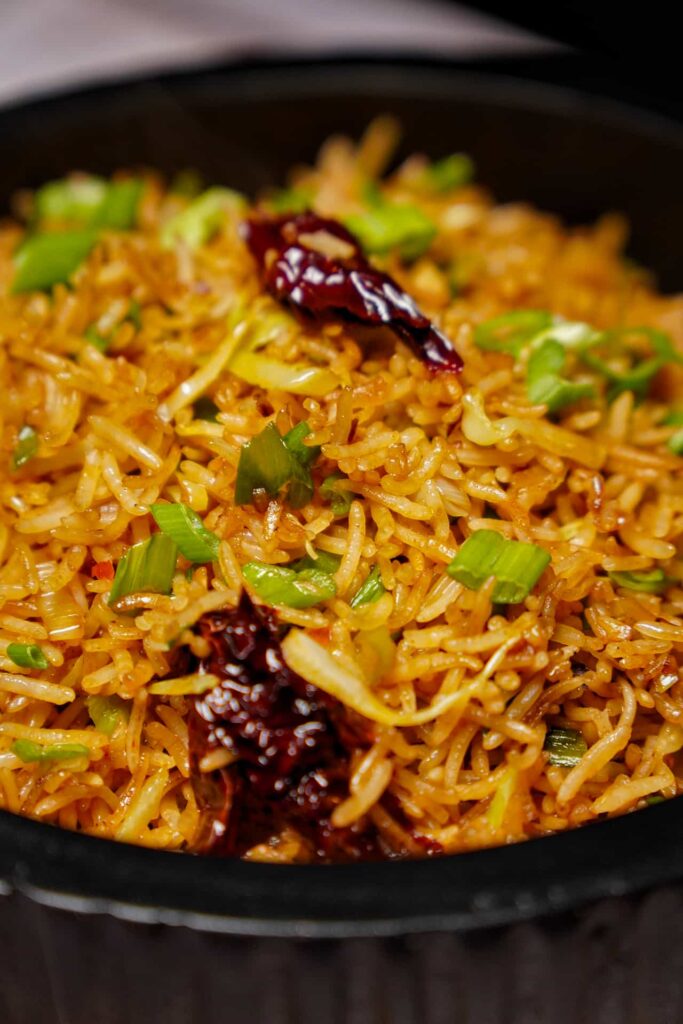An extreme close-up of Vegetable Schezwan Fried Rice, showing the detailed texture of the long-grain rice, chopped scallions, and spicy red chili peppers.