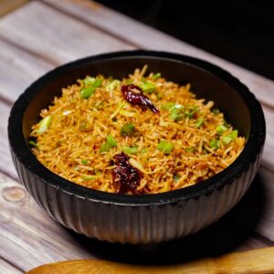 An eye-level view of a bowl of Schezwan fried rice on a rustic wooden table next to a wooden spatula, highlighting the vibrant colors and garnishes.