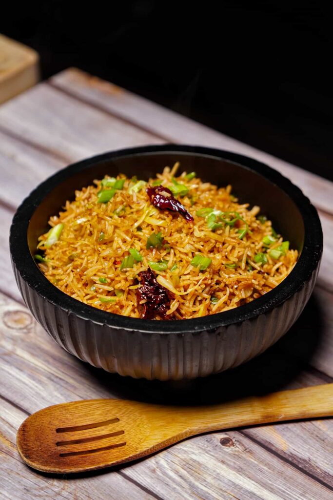 An eye-level view of a bowl of Schezwan fried rice on a rustic wooden table next to a wooden spatula, highlighting the vibrant colors and garnishes.