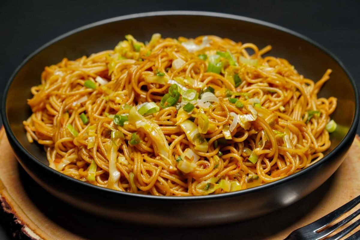 An angled eye-level shot of vegetable noodles in a matte black bowl resting on a rustic wood slice, with a black fork visible in the corner.