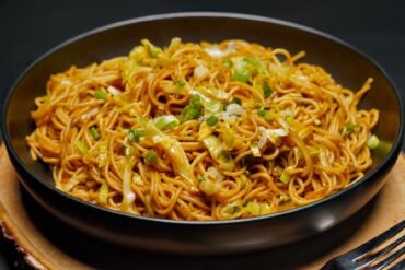 An angled eye-level shot of vegetable noodles in a matte black bowl resting on a rustic wood slice, with a black fork visible in the corner.