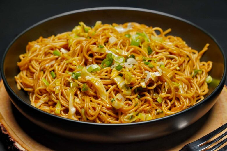 An angled eye-level shot of vegetable noodles in a matte black bowl resting on a rustic wood slice, with a black fork visible in the corner.