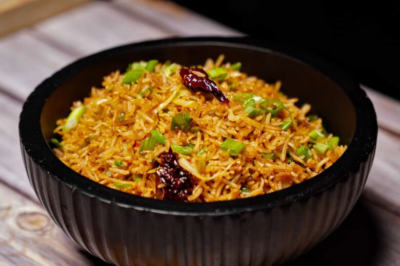 A close-up, slightly angled shot of spicy Vegetable Schezwan Fried Rice in a black fluted bowl, featuring golden rice, green onions, and dried red chilies.