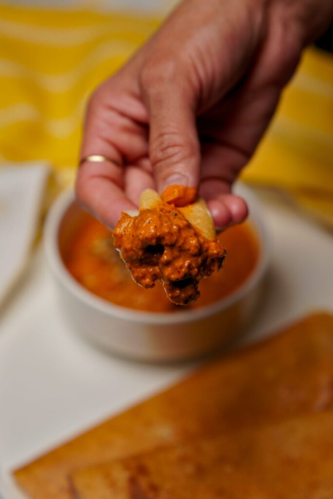 A hand dipping a piece of crispy golden dosa into a bowl of thick, orange-red tomato chutney.