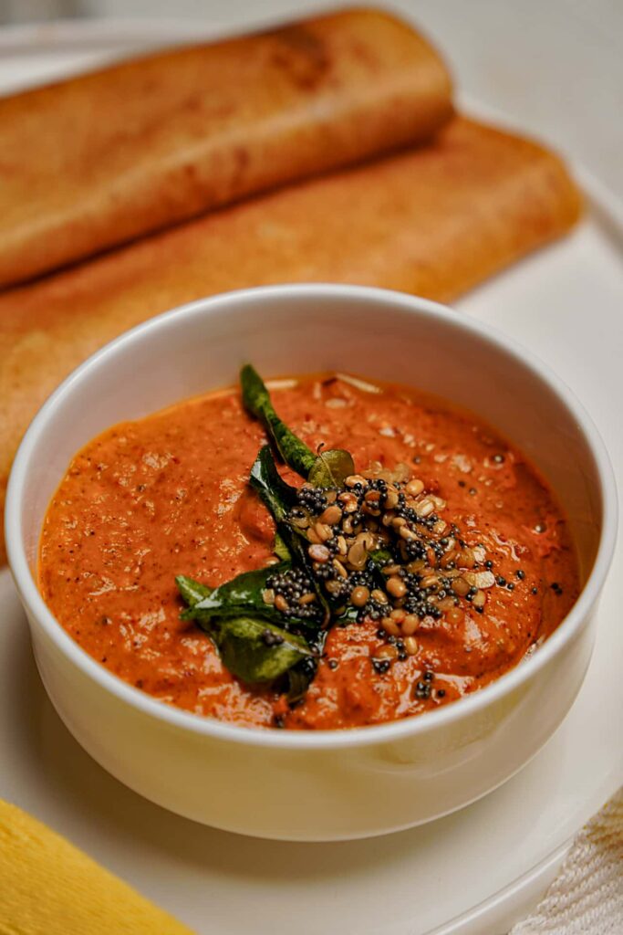A white bowl filled with spicy tomato chutney topped with mustard seed and curry leaf tempering, served with folded crispy dosas on the side.