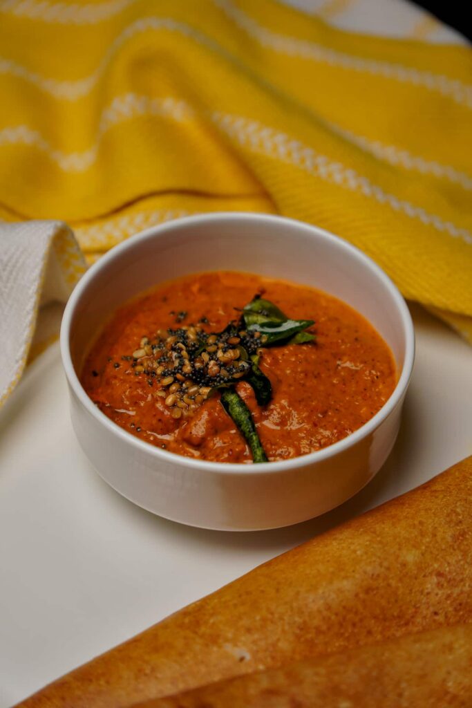 A white bowl filled with spicy tomato chutney topped with mustard seed and curry leaf tempering, served with folded crispy dosas on the side.