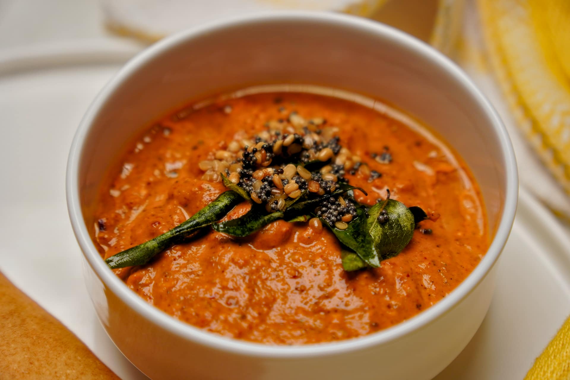 Macro view of a bowl of tomato chutney showing the texture of the blended tomatoes and the tempering of roasted lentils and mustard seeds on top.