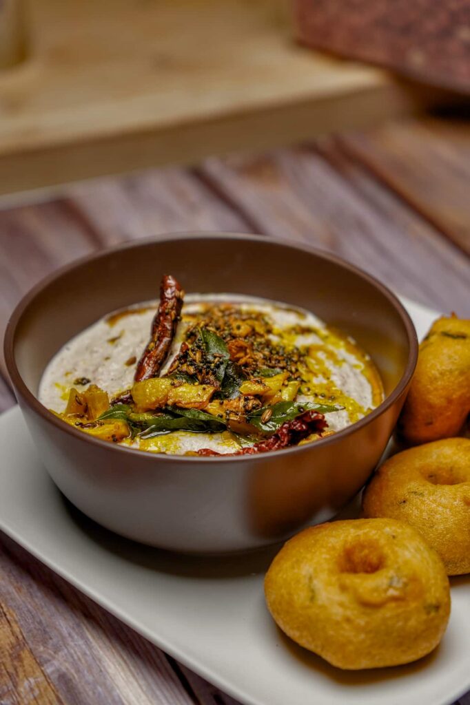 A dark brown bowl of creamy peanut chutney topped with aromatic tempering, served alongside a stack of golden-brown fried medu vadas on a white plate.