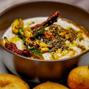 Vertical shot of a bowl of peanut chutney with a generous drizzle of oil and spices, with medu vadas blurred in the foreground.