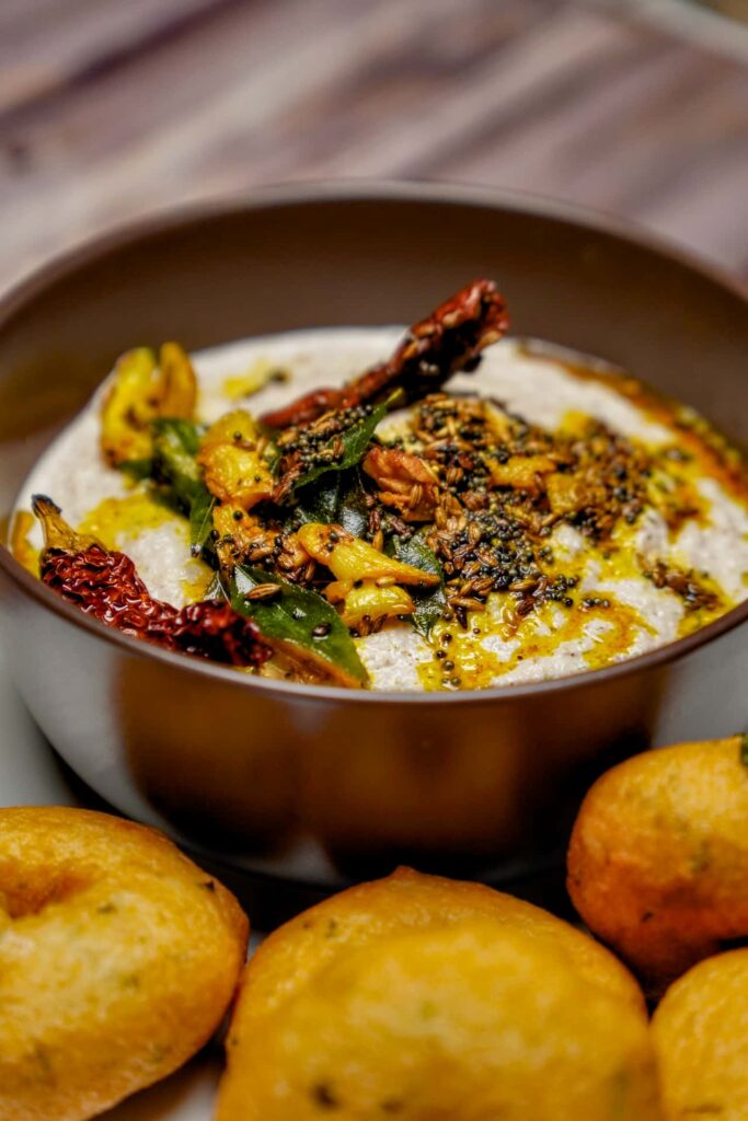 Vertical shot of a bowl of peanut chutney with a generous drizzle of oil and spices, with medu vadas blurred in the foreground.