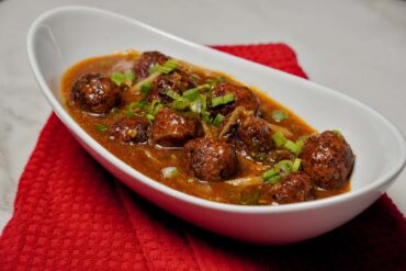 A white bowl filled with glossy, dark red Veg Manchurian gravy topped with fresh green spring onions, served on a white bowl.