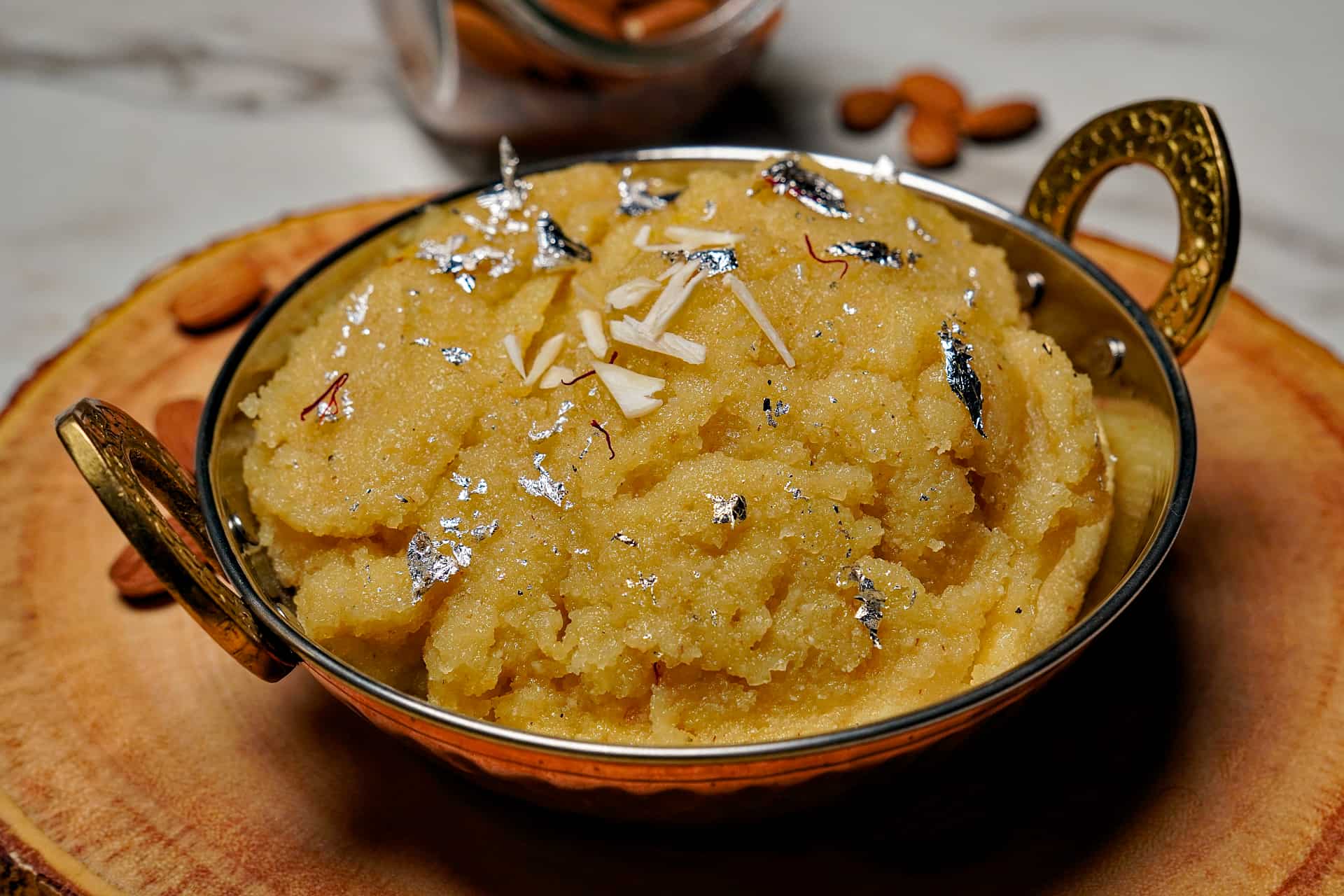 A wide shot of rich, yellow Badam Halwa in a traditional bowl, garnished with sliced almonds and green onions on a wooden board.