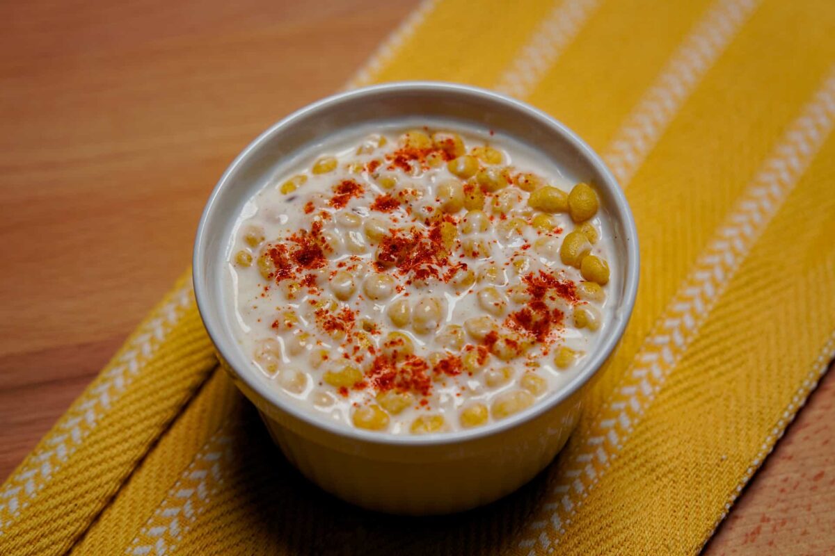 A wide shot of creamy Boondi Raita in a white bowl, topped with golden boondi and red chili.