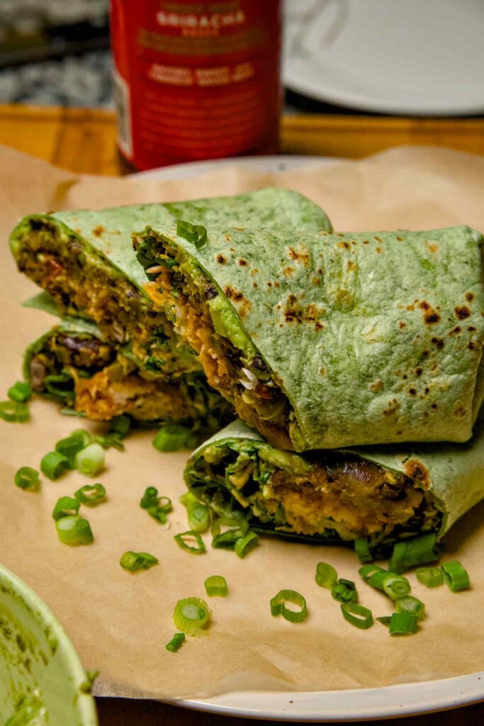 Two halves of a spinach wrap breakfast burrito topped with green onions, with a bottle of Sriracha sauce.