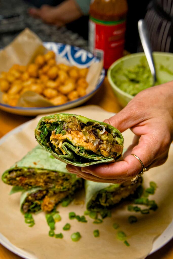 A hand holding a cross-section of a vegetarian breakfast burrito, featuring avocado, greens, and tater tots inside a green tortilla.