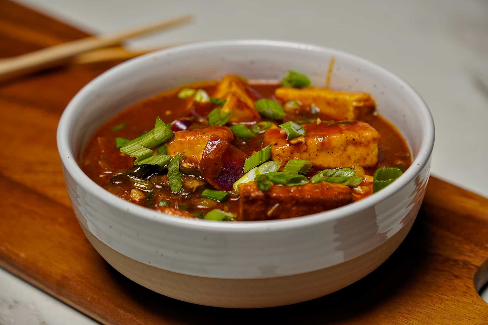 medium shot of a white ceramic bowl filled with Chilli Paneer gravy, featuring pan-fried paneer cubes, red onions, and a garnish of fresh green onions on a wooden board.