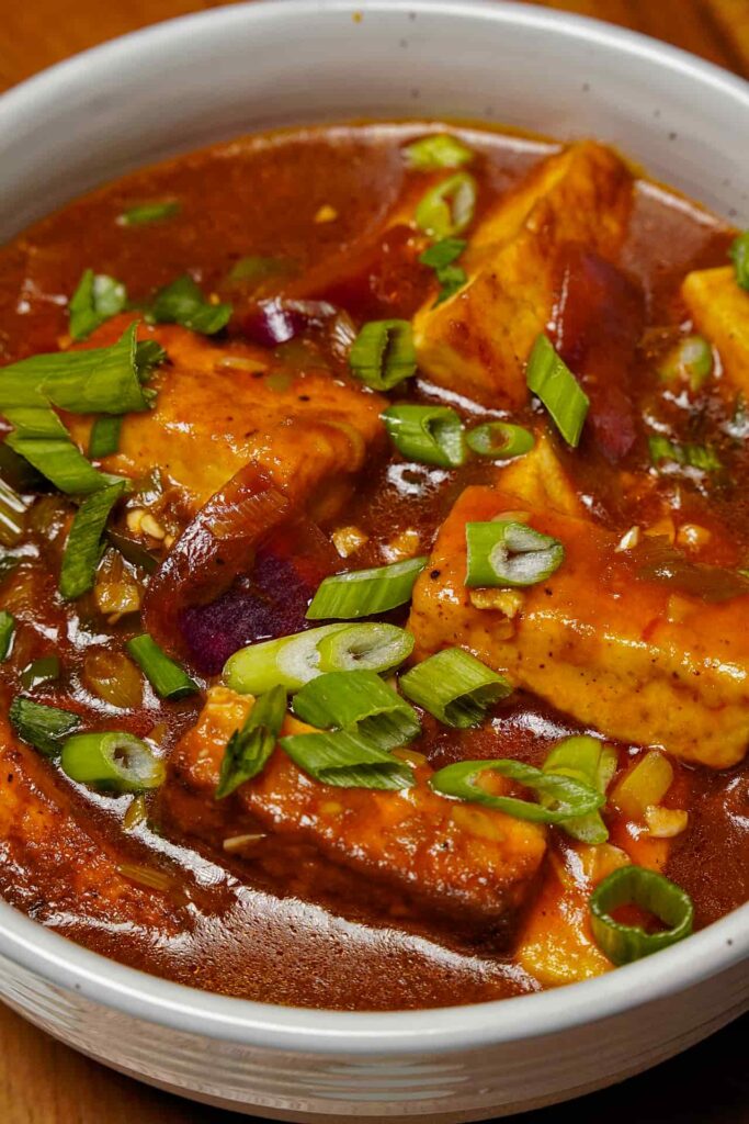 A detailed top-down view of the Chilli Paneer dish, showing the texture of the spicy gravy, cooked onions, and the fresh green garnish.