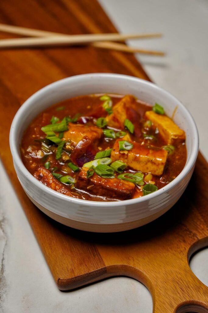 A slightly high-angle view of a bowl of Chilli Paneer on a wooden serving board with wooden chopsticks resting in the blurred background.