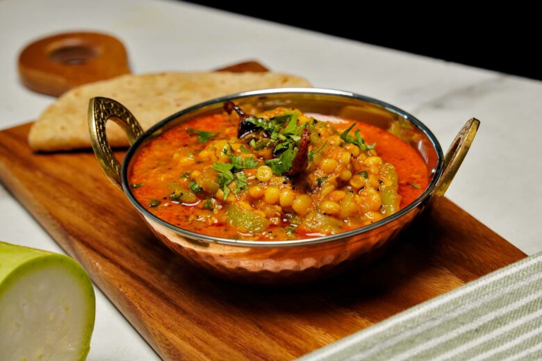 A copper bowl of Lauki Chana Dal garnished with fresh cilantro and dried red chilis, served with flatbread on a wooden board.