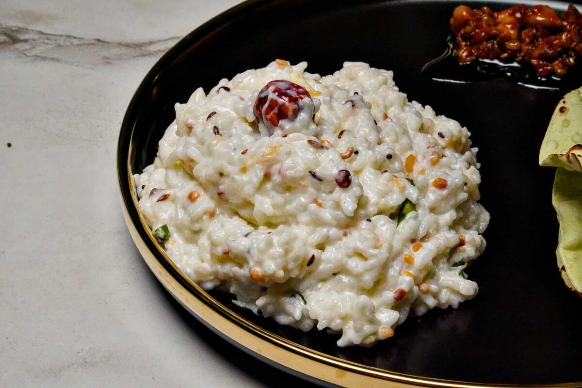 A medium shot of a plate filled with creamy Curd Rice, garnished and served on with Papad and Pickle.