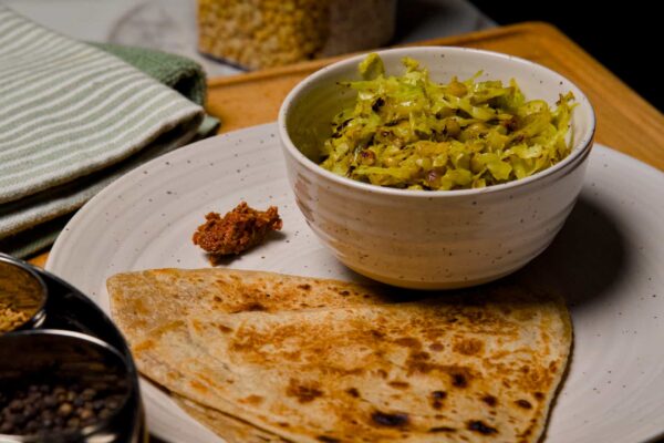 A white bowl filled with cabbage chana dal sabzi served on a plate with folded wheat parathas and a small portion of red pickle.