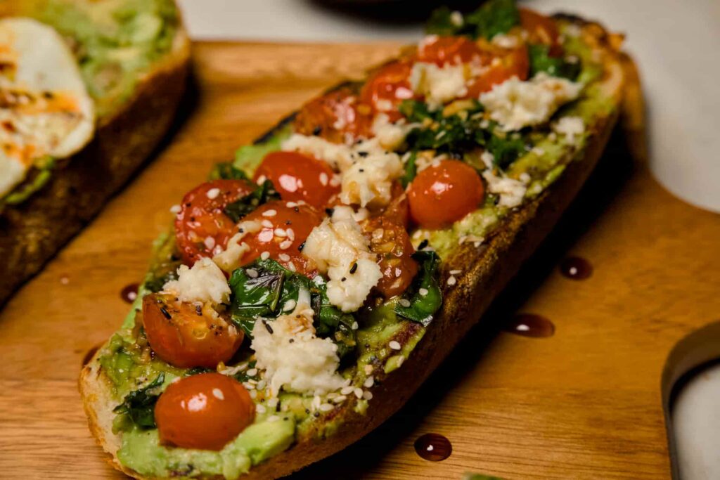 A side profile of avocado toast topped with halved cherry tomatoes, fresh mozzarella pearls, torn basil, and sesame seeds.