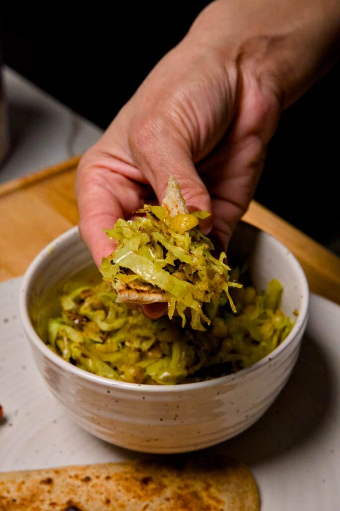 A hand holding a piece of wheat paratha scooped with a generous portion of cabbage and chana dal sabzi over the bowl.