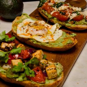 An overhead diagonal view of three different avocado toast recipes—tofu, egg, and caprese—arranged on a wooden serving platter.