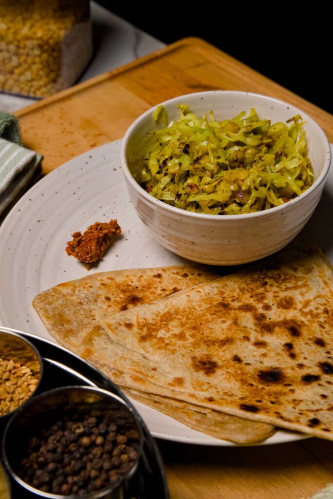 Overhead view of a complete meal featuring cabbage chana dal sabzi, parathas, and a small spice container on a wooden board.