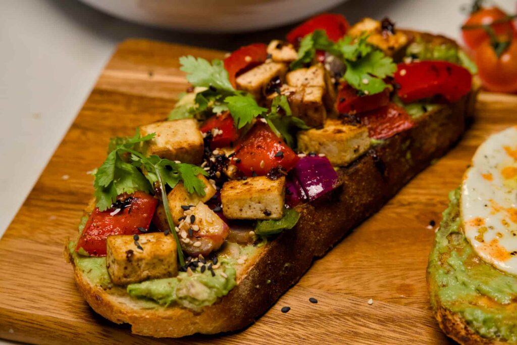 Close-up of a vegan avocado toast variation topped with sautéed spicy tofu cubes, red bell peppers, red onions, and fresh cilantro.