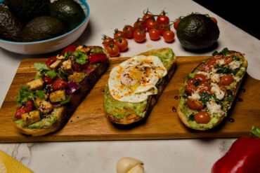 Three slices of gourmet avocado toast on a wooden serving board featuring egg, caprese, and spicy tofu toppings, surrounded by fresh tomatoes and avocados.