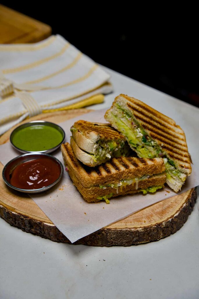 A close-up of a sliced Mumbai Grill Sandwich served on a wooden board with small bowls of red tomato ketchup and green coriander chutney.