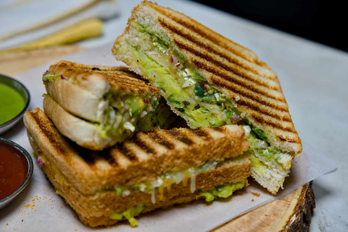 A close-up of a sliced Bombay Grill Sandwich served on a wooden board with small bowls of red tomato ketchup and green coriander chutney.