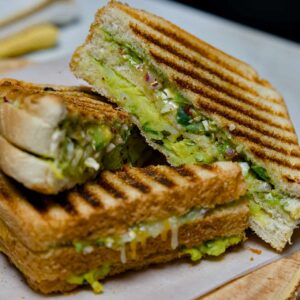 A close-up of a sliced Bombay Grill Sandwich served on a wooden board with small bowls of red tomato ketchup and green coriander chutney.