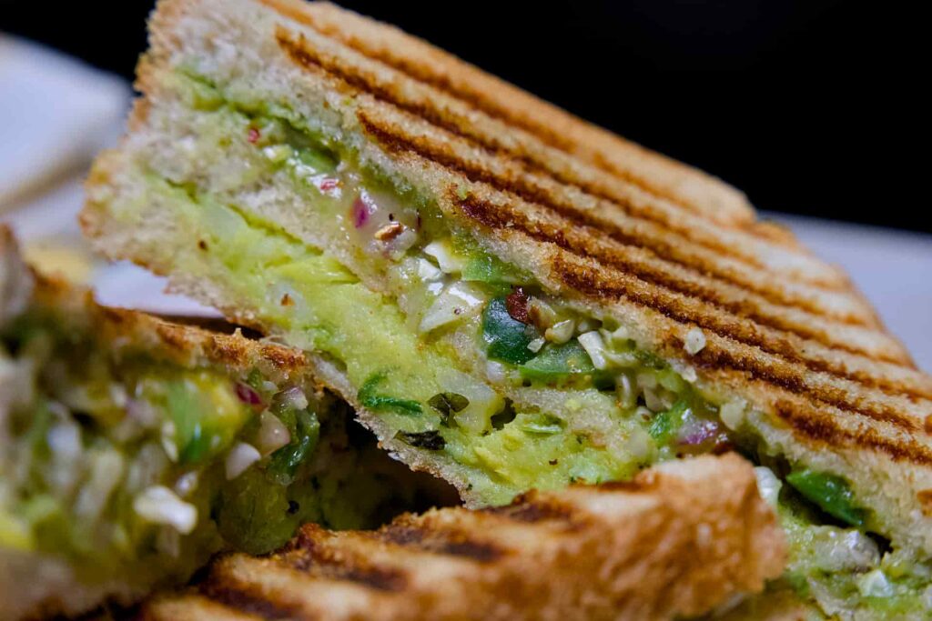 Macro shot of the Bombay Grill Sandwich cross-section showing the vibrant green chutney, mashed potato layer, and melted mozzarella cheese.