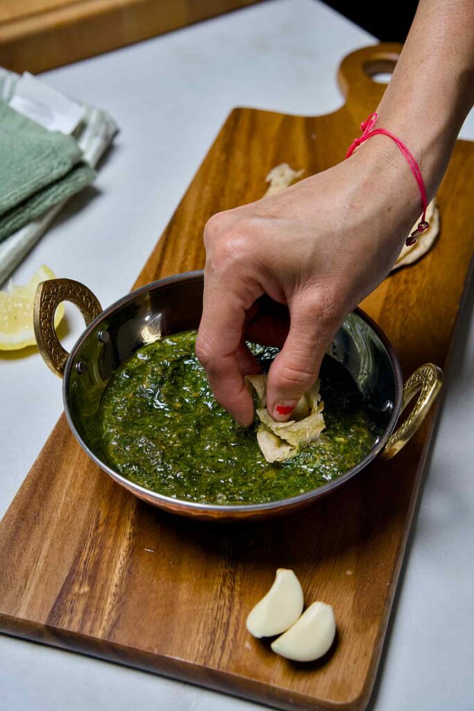 A hand scooping a bite of vibrant green Suva ni Bhaji (Gujarati Dill Curry) with a piece of warm chapati from a traditional copper serving bowl.